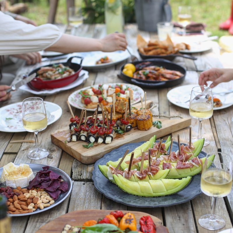People eating food with wine on a wooden table outside.