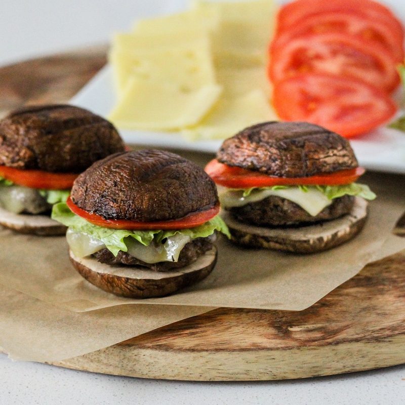 Three mushroom bun sliders with cheese, tomatoes, and lettuce on a rectangular plate on a circular wooden tray.