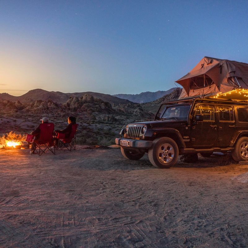 A couple camping by a campfire with a jeep with a rooftop tent.