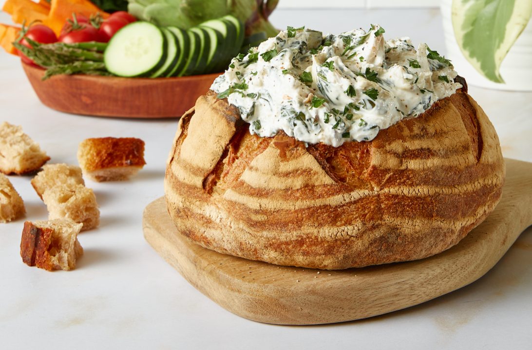 Good Culture Spinach and Artichoke dip placed in a bread bowl on a white kitchen counter with salad in the back.