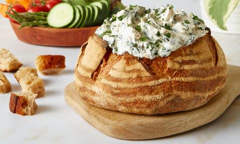 Good Culture Spinach and Artichoke dip placed in a bread bowl on a white kitchen counter with salad in the back.