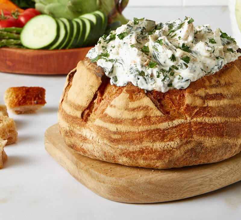 Good Culture Spinach and Artichoke dip placed in a bread bowl on a white kitchen counter with salad in the back.
