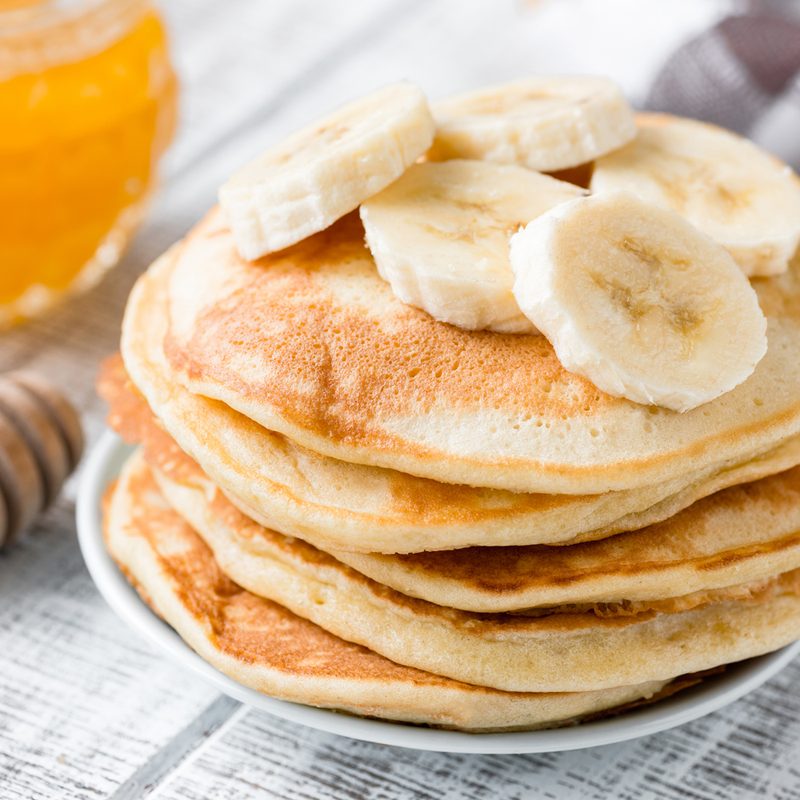 Banana slices on a plate of pancakes beside a cup of honey on a table.