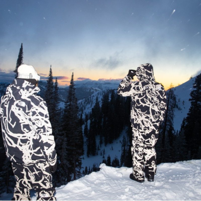 Two people wearing a matching winter coats and snow pants from The North Face XX Kaws collection.