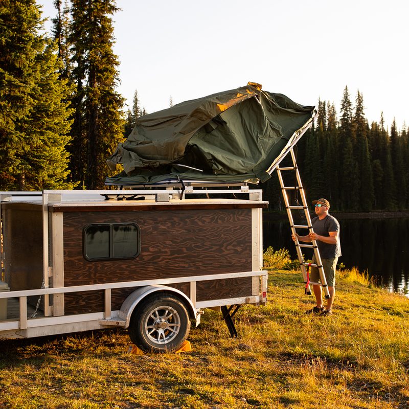 A man climbing a ladder to set up a rooftop tent on top of his vehicle at a waterfront campsite.