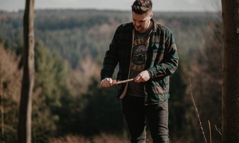 A man in a green and gray jacket whittles a stick to a sharp point