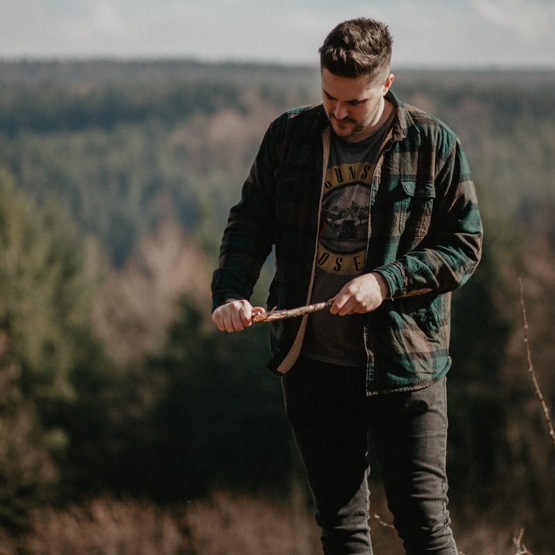 A man in a green and gray jacket whittles a stick to a sharp point