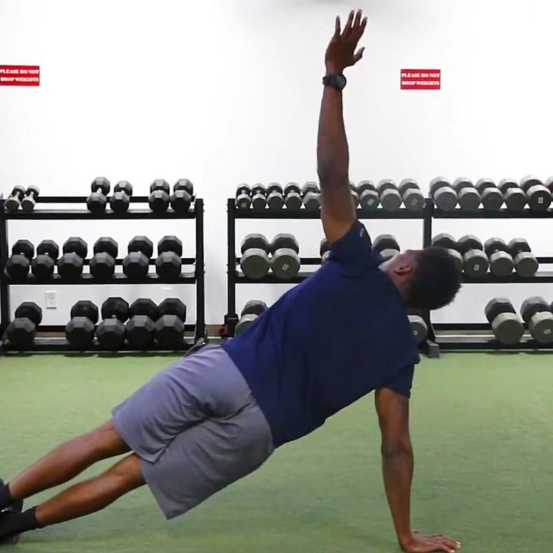 A male athlete doing thoracic rotations with dumbbells on racks in a gym.