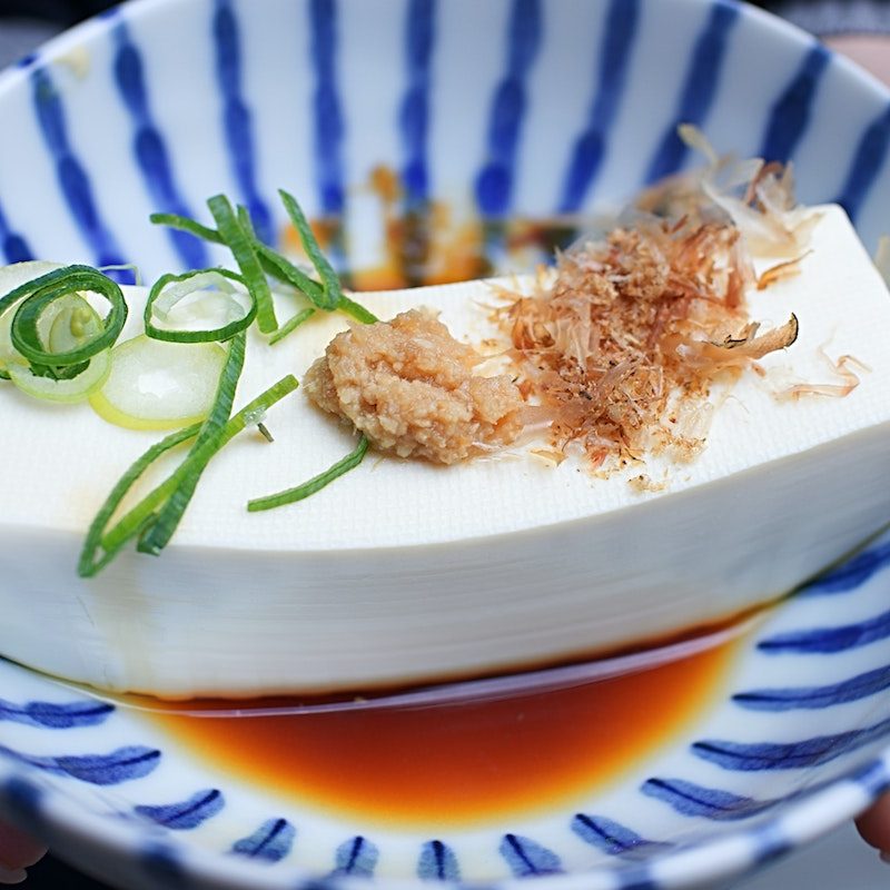 Block of tofu in a bowl with broth and garnishes.