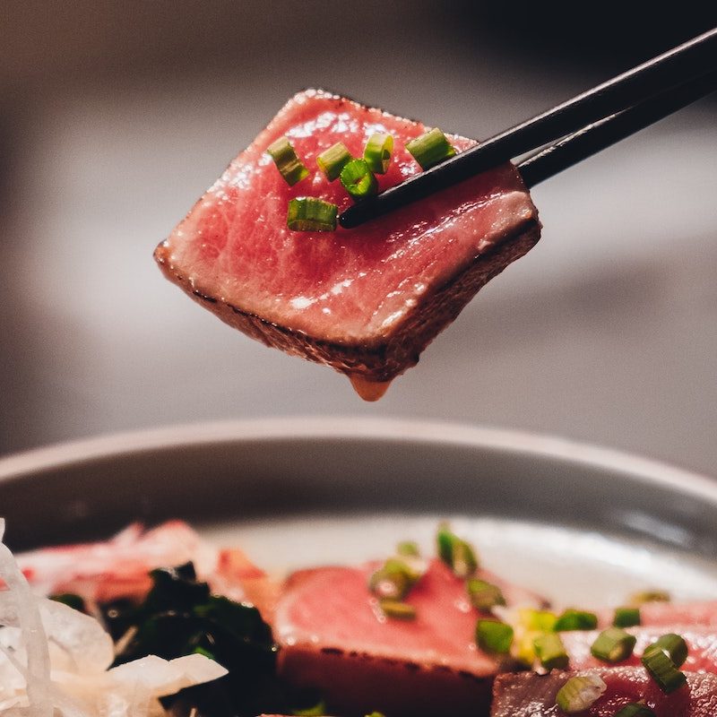A slice of seared tuna with garnish of chives held in chopsticks.