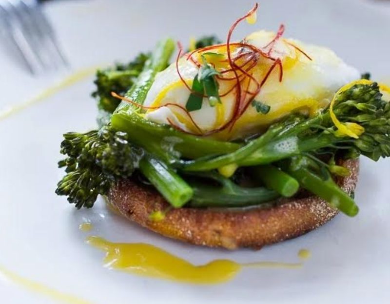 One eggs Benedict with broccolini on white plate with fork.
