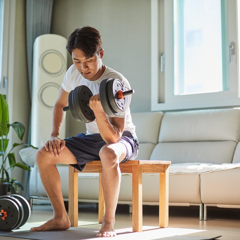 A man sitting on a table while lifting a dumbbell in the living room.