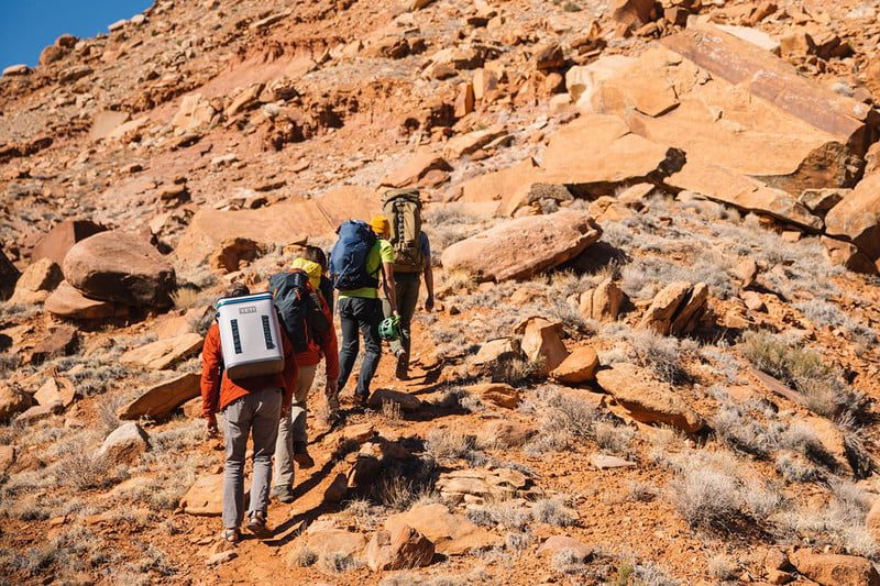 Four people hiking on a rocky trail. The last member is carrying a Yeti backpack cooler.