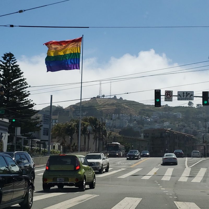 An LGBTQ+ flag and a view of Twin Peaks and the Sutro Tower from the Castro District in San Francisco, California.
