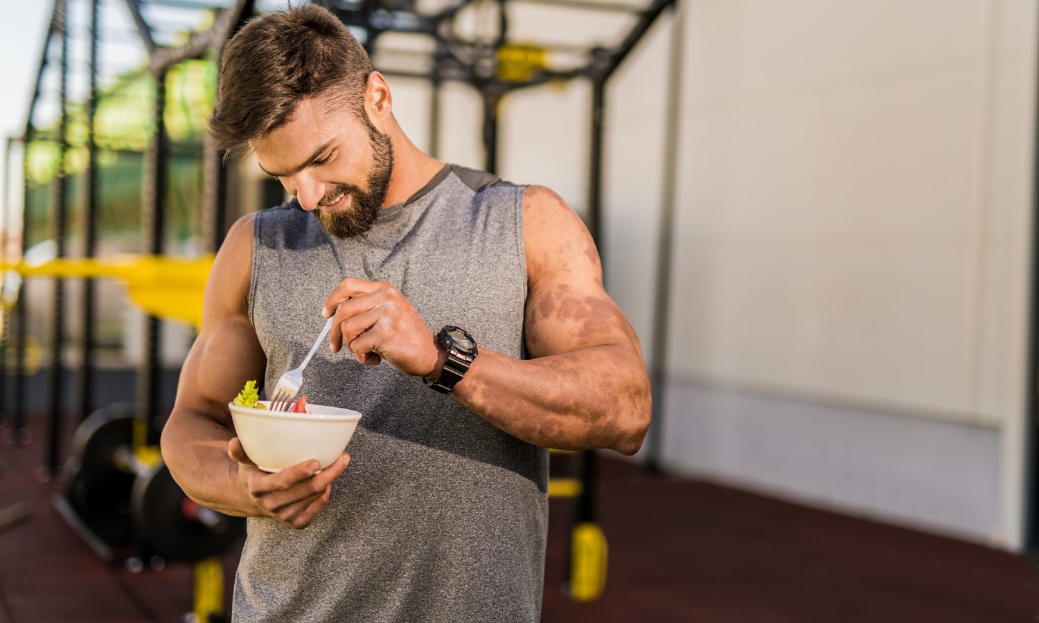 A man in gray sleeveless top eating a vegetable salad in the gym.