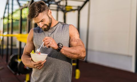 A man in gray sleeveless top eating a vegetable salad in the gym.