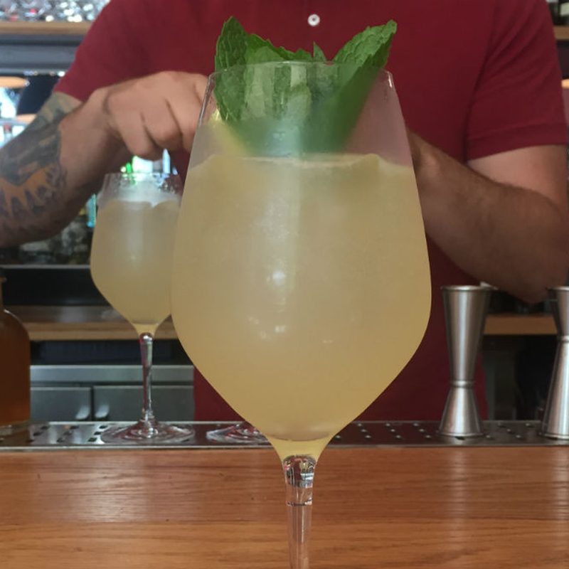 A male bartender standing behind a bar counter with a glass of Almond and Peach Spritz with mint sprig garnish