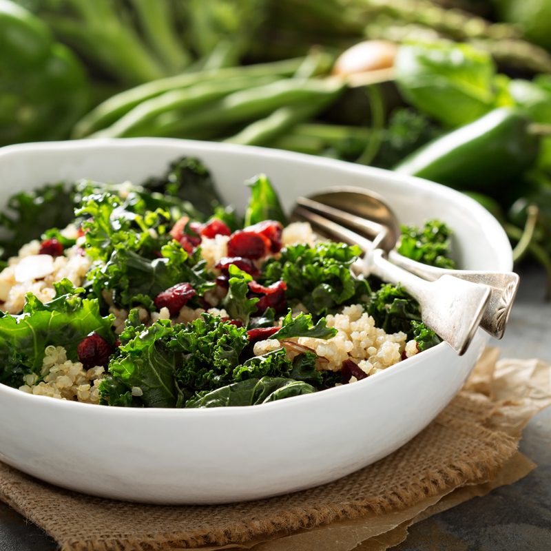 A bow of kale and quinoa salad on brown cloth with vegetables in the background.