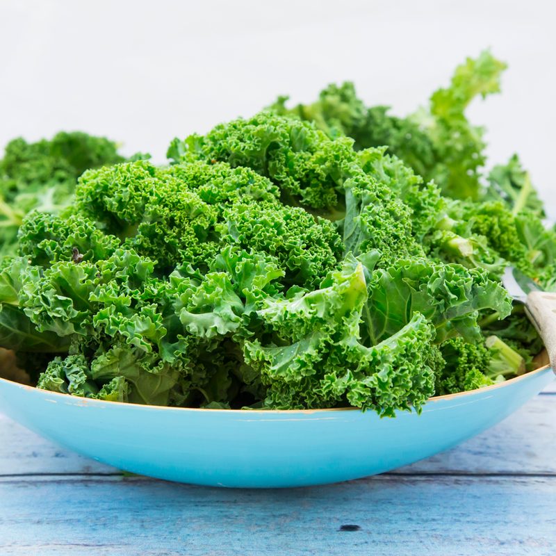 Fresh kale leaves and a knife on a white bowl on a white wooden table.
