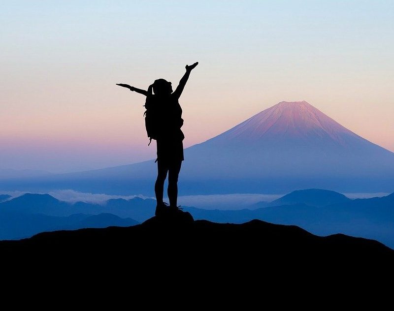 A backpacker on top of a mountain, raising her arms in a "Y" shape.