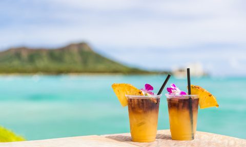 Two Mai Tai cocktails topped with flowers and pineapples on a surface with a view of the beach