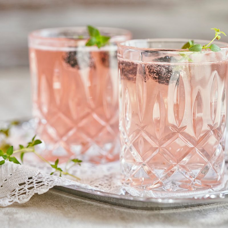 Two glasses of Rose cocktails with blueberry garnish on crocheted cloth on a metal tray