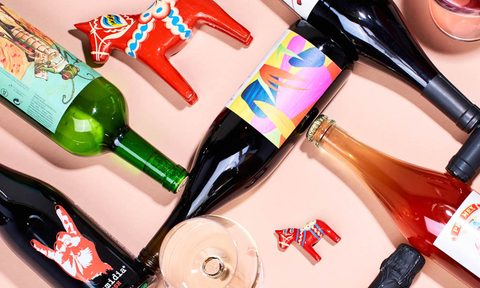 An overhead shot of an empty wine glass, a glass filled with wine, wine bottles, and differently sized red wooden horses on pink table.