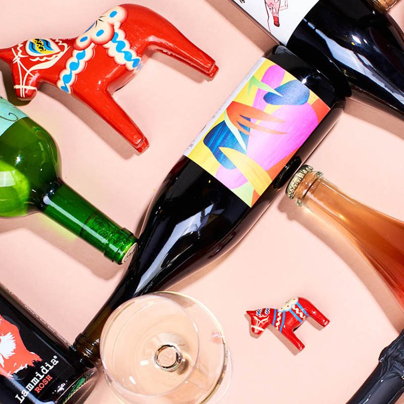 An overhead shot of an empty wine glass, a glass filled with wine, wine bottles, and differently sized red wooden horses on pink table.