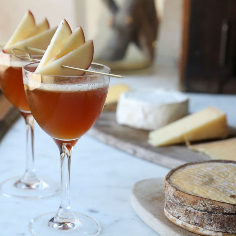 Two glasses of Between the Apple Trees cocktails garnished with apple slices beside cheeses on oval-shaped wooden plates.