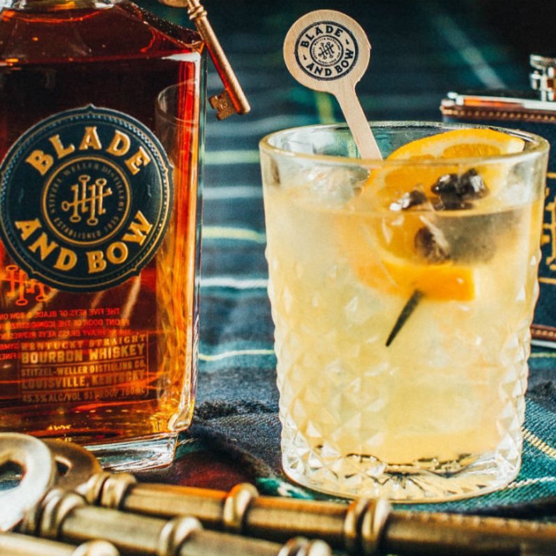 A glass of Blade and Bow New Fashioned cocktail with orange and blueberry skewer garnish beside a bottle of Kentucky Straight Bourbon Whiskey.