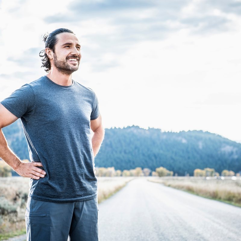 A man with his hands on his hips standing on an empty road with a smile.