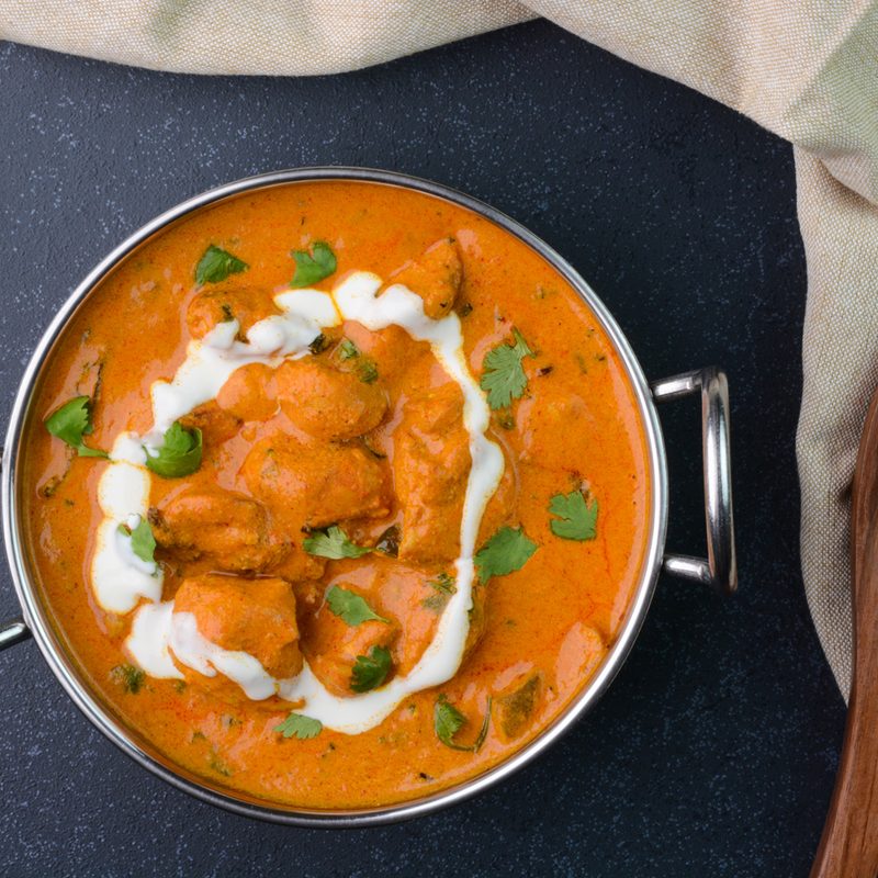 An overhead shot of butter chicken in a pot beside herbs and a wooden spoon.