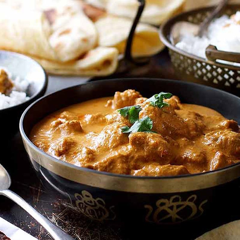 A butter chicken served in a bowl beside a metal spoon with various dishes in the background.