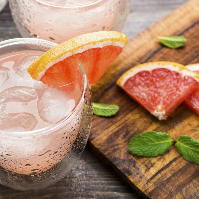 A glass of Cancion del Verano garnished with grapefruit beside ice cubes, grapefruit wedges, leaves, and a handle of a knife on wooden cutting board