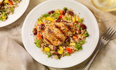 An overhead shot of grilled chicken with rice and vegetables beside a metal fork and a glass of wine on a table.
