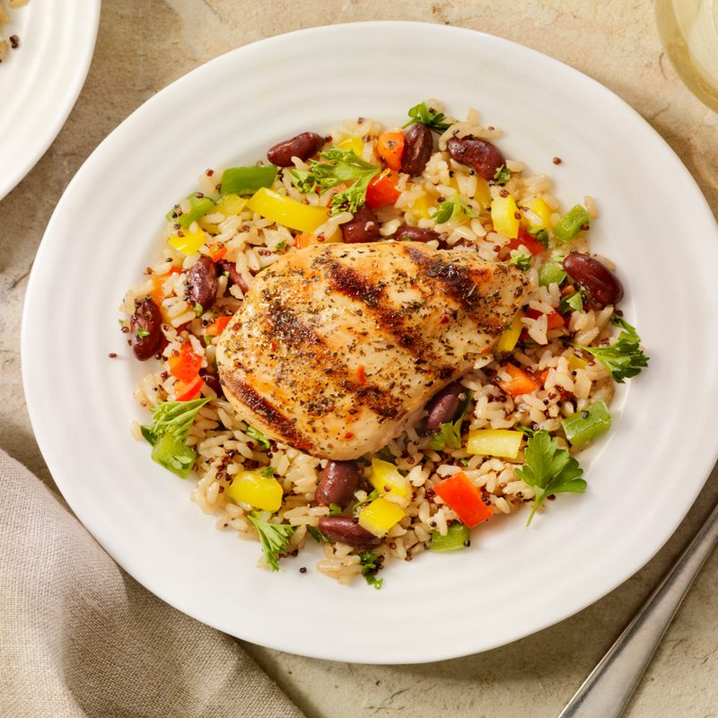 An overhead shot of grilled chicken with rice and vegetables beside a metal fork and a glass of juice on a table.