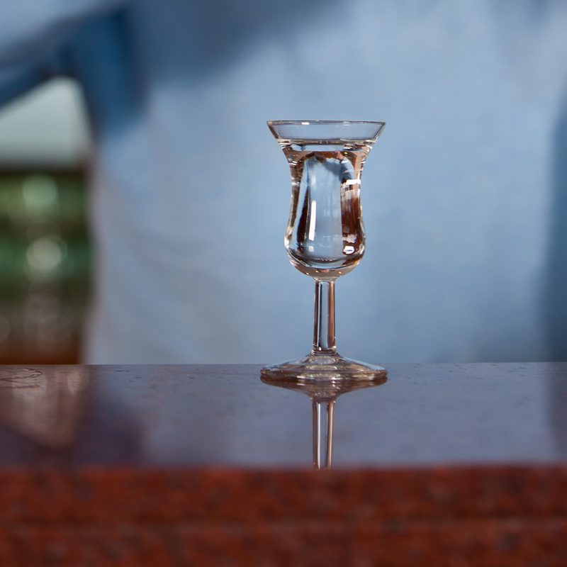 A glass of genever on bar counter with a midsection of a man behind it.