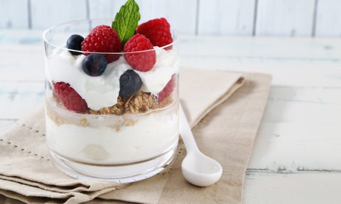 A glass of yogurt parfait with berries and granola beside a plastic spoon on brown cloth