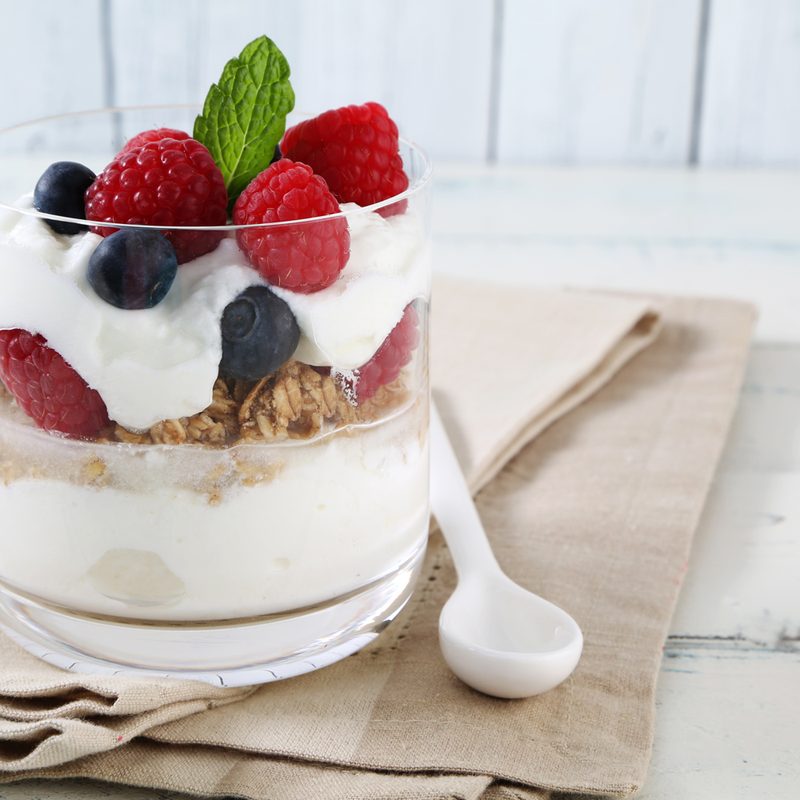 A glass of yogurt parfait with berries and granola beside a plastic spoon on brown cloth.