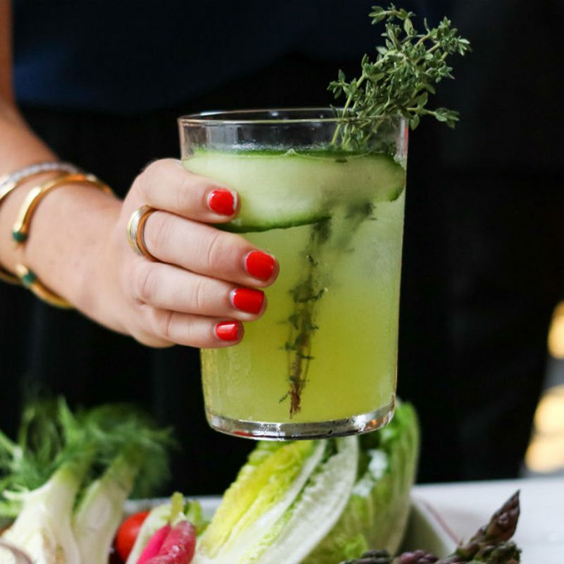 A woman holds a glass of Greenmarket Spritz containing cucumber slices and thyme sprig over vegetables