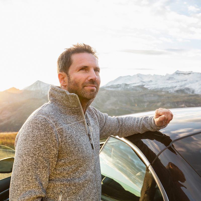 A man in gray half-zip pullover sweater placing his arm on his car in the outdoors.