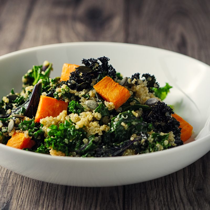 A bowl of kale salad with roasted sweet potatoes and pumpkin seeds beside a fork on a wooden table.