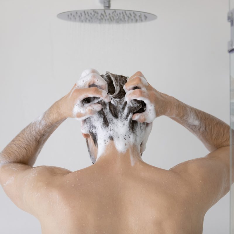 A rear view of a man lathering shampoo on his hair in the shower.