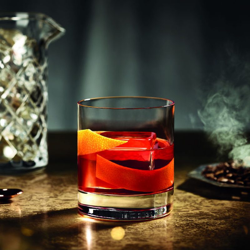 A Jitney Negroni garnished with orange on a table with spoon, pitcher with ice, and cigarette tray in the background.