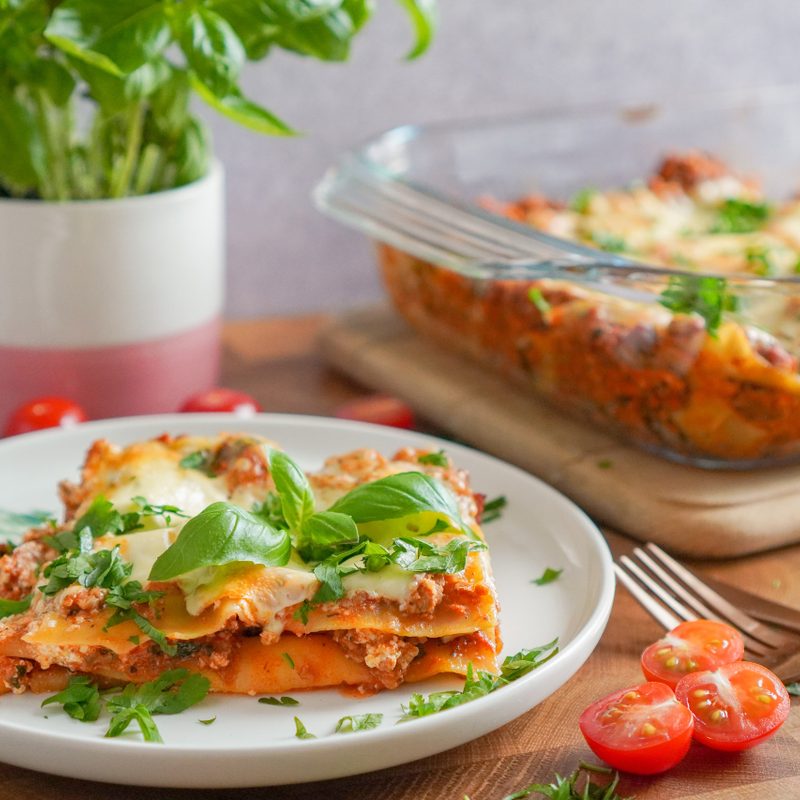 Slice of lasagna in a white plate with basil and lasagna dish in background