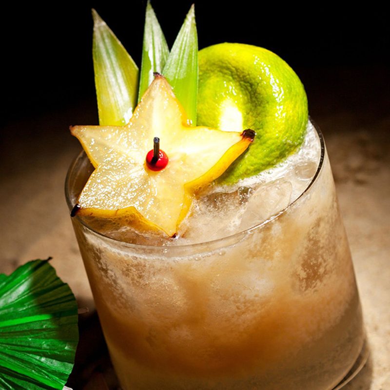 A Mai Tai cocktail topped with fruits and leaves beside a cocktail umbrella