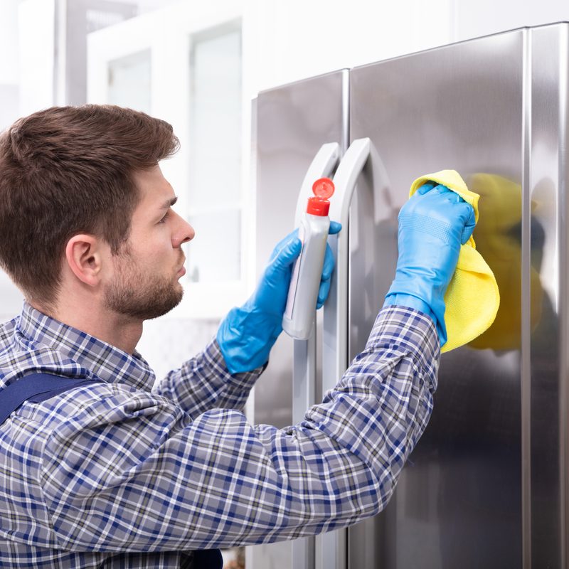 Side View Of Man Cleaning Refrigerator With Cloth And Bottle.