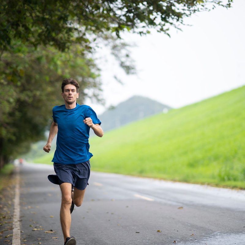A man in blue shirt and navy shorts jogging on a road with trees and grassy field in the background.