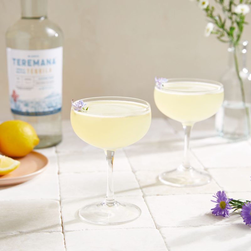 Two glasses of Mana Daisy cocktail with an empty bottle of liquor, a plate of lemons, purple flowers, and white flowers in a bottle
