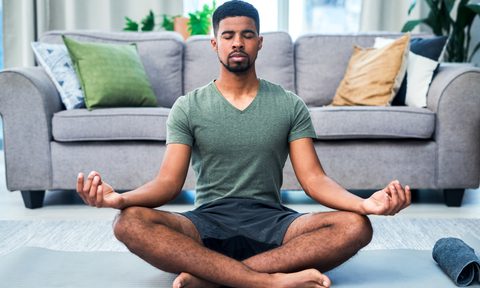 A man in a green shirt and gray shorts meditating on a yoga mat in the living room.